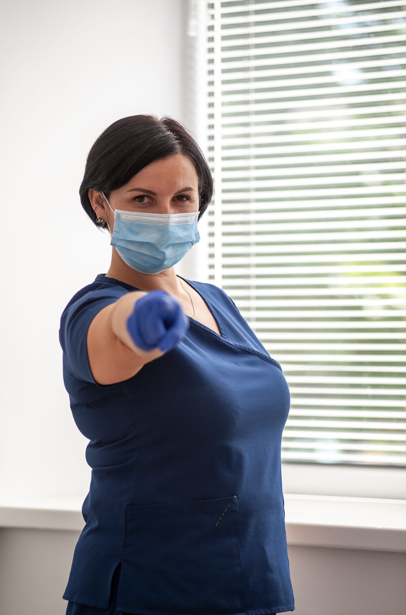 Woman in blue scrubs with mask and gloves pointing towards camera indoors.